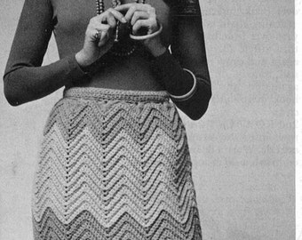 Peut inclure: Une photo en noir et blanc d'une femme portant un pull noir à col roulé à manches longues et une jupe en crochet avec un motif en chevron. Elle porte un collier et a les cheveux longs. Elle est debout, les jambes légèrement écartées et les bras croisés devant elle.