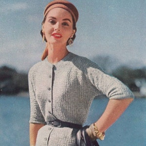 May include: A woman wearing a light gray knitted dress with a brown hat, a black belt, and a black handbag. She is standing in front of a body of water.
