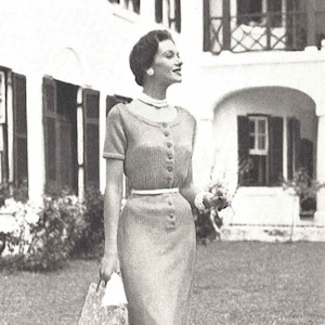 May include: A woman in a gray knitted dress with a white belt and short sleeves. She is carrying a white mesh bag and walking on a grassy lawn.