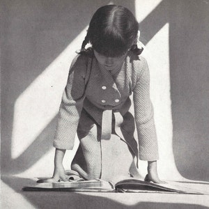 Könnte beinhalten: Ein junges Mädchen in einem weißen Strickmantel mit Knöpfen und Gürtel, kniet und liest ein Buch.