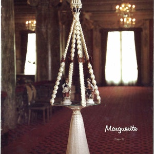 Peut inclure: Une suspension en macram&eacute; blanche avec un pompon, orn&eacute;e d'un sommet d&eacute;coratif et de deux petites figurines. La suspension est pr&eacute;sent&eacute;e dans une pi&egrave;ce avec un tapis rouge et un lustre.