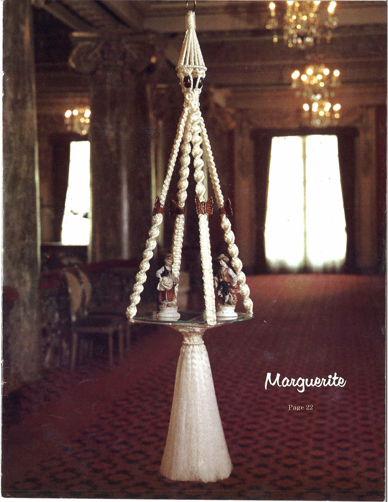Peut inclure: Une suspension en macram&eacute; blanche avec un pompon, orn&eacute;e d'un sommet d&eacute;coratif et de deux petites figurines. La suspension est pr&eacute;sent&eacute;e dans une pi&egrave;ce avec un tapis rouge et un lustre.