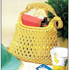 May include: A yellow crocheted tote bag with a yellow plastic handle and yellow beads around the bottom. The bag is filled with items like a pink towel, a bottle of sunscreen, and a small white and yellow patterned bag. The bag is sitting on a sandy surface with a blue plastic shovel and a white seashell.