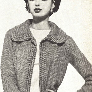 May include: A close-up of a woman wearing a grey knitted cardigan with a white collar and a white shirt underneath. The cardigan has a textured design and a button closure. The woman is wearing a black skirt and has her hair styled in a bouffant.