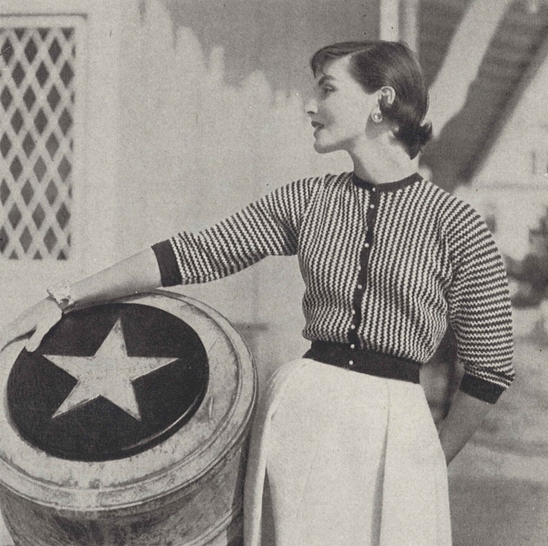 May include: A black and white striped cardigan with a button closure. The cardigan is worn over a white skirt. The model is leaning against a barrel with a white star on the top.