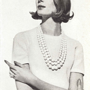 Puede incluir: Una mujer con un suéter blanco y un collar de perlas. Tiene el pelo corto y oscuro y lleva un suéter blanco sencillo con un collar de perlas. Tiene los brazos cruzados y mira hacia un lado.