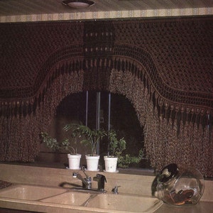 May include: A brown macrame curtain with fringe hangs over a window in a kitchen. The curtain is arched in the center and has a geometric pattern. There are three potted plants on the windowsill below the curtain.