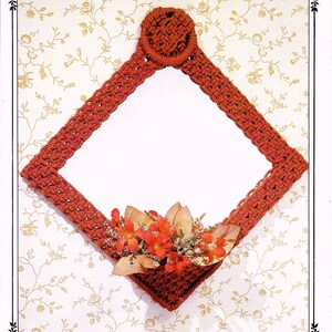 以下が含まれることがあります： 花柄のダイヤモンド型のクロシェ編み壁掛け。赤い糸で編まれており、上部に小さな円形があります。壁掛けの上には「Floral Reflections」というタイトルが印刷されています。