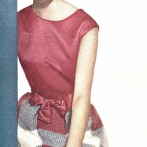 May include: A woman wearing a pink and white checkered dress with a pink bow at the waist. She is holding a blue wall with her left hand and has a ring on her right hand.