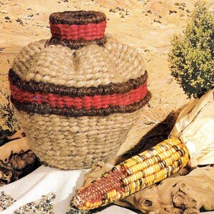 May include: A woven brown and red rope vase with a corn cob with husks on a wooden surface.