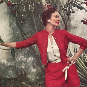 May include: A red knitted cardigan with a white knitted top underneath. The cardigan has a button closure and the top has a V-neck. The woman is wearing a red skirt and white bracelets.