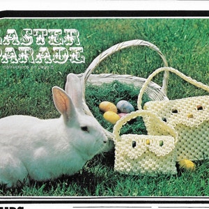 Puede incluir: Un conejo blanco se sienta en la hierba junto a una cesta de mimbre llena de huevos de Pascua. Dos bolsas de ganchillo, una grande y otra pequeña, están delante de la cesta. Las bolsas son blancas y tienen un borde festoneado. El texto "Easter Parade" está en la parte superior de la imagen con las palabras "instructions on page 8" debajo.