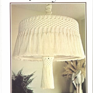 May include: A white macrame lampshade with a fringe border and a tassel hanging from the bottom. The lampshade is hanging from a ceiling fixture.