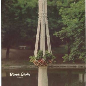 May include: A white macrame hanging planter with a red pot filled with green plants. The planter has a decorative top and a long, fringed bottom. The text "Bonne Santé" is visible.