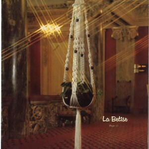 Peut inclure: Un porte-plante en macram&eacute; blanc avec des perles en bois et un pot noir contenant une plante verte. Le porte-plante est suspendu au plafond dans une pi&egrave;ce avec un tapis rouge et des murs orn&eacute;s. Le texte "La Betise Page 13" est visible dans le coin inf&eacute;rieur droit de l'image.