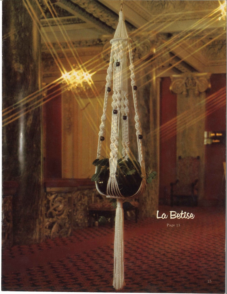 Peut inclure: Un porte-plante en macram&eacute; blanc avec des perles en bois et un pot noir contenant une plante verte. Le porte-plante est suspendu au plafond dans une pi&egrave;ce avec un tapis rouge et des murs orn&eacute;s. Le texte "La Betise Page 13" est visible dans le coin inf&eacute;rieur droit de l'image.