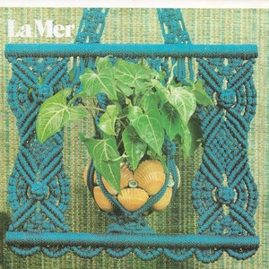 May include: A teal macrame plant holder with a seashell pot holding a green leafy plant. The macrame design features intricate knots and a shelf at the bottom. The text "La Mer" is at the top.