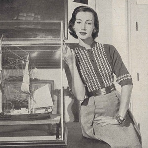Puede incluir: Una mujer con un suéter de patrón blanco y negro con cinturón y falda, de pie junto a una vitrina con un modelo de barco en su interior.