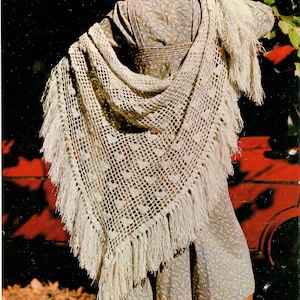 Puede incluir: Un chal blanco de ganchillo con flecos, usado sobre un vestido de estampado floral. El chal tiene un delicado patrón de calado y está drapeado sobre los hombros de una persona que lleva botas marrones.