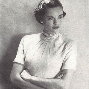 May include: A woman wearing a white knitted sweater with a high neckline and short sleeves. She is wearing a pearl necklace and has her hair styled in a short, curled updo.