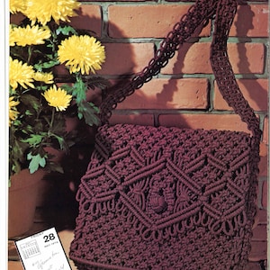 Może przedstawiać: Brązowa torebka wykonana techniką makramy z geometrycznym wzorem. Torebka ma długi pasek i wisi na ceglanej ścianie. Na pierwszym planie znajdują się żółte kwiaty w doniczce.