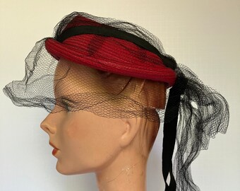 May include: A red hat with a black ribbon and a black net veil. The hat is displayed on a mannequin head. The hat is a vintage style.