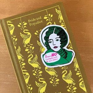 Puede incluir: Un libro de tapa dura de "Orgullo y Prejuicio" de Jane Austen, con una cubierta dorada con un patr&oacute;n de cisnes. Una pegatina con una ilustraci&oacute;n verde y blanca de una mujer y el texto "I send NO compliments to your mother" est&aacute; en el libro.