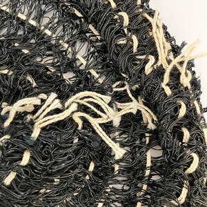 May include: Close-up of a woven textile with a textured appearance. The piece is primarily black with interwoven cream-colored threads creating a grid-like pattern. The edges have loose, frayed threads.