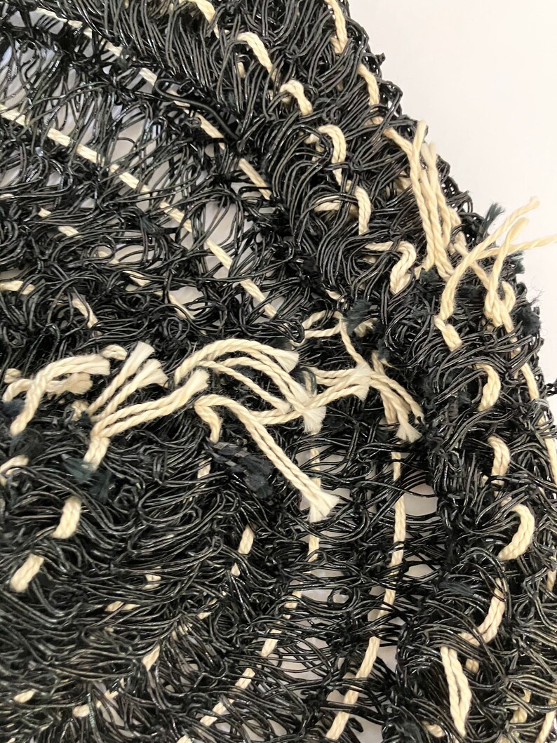 May include: Close-up of a woven textile with a textured appearance. The piece is primarily black with interwoven cream-colored threads creating a grid-like pattern. The edges have loose, frayed threads.