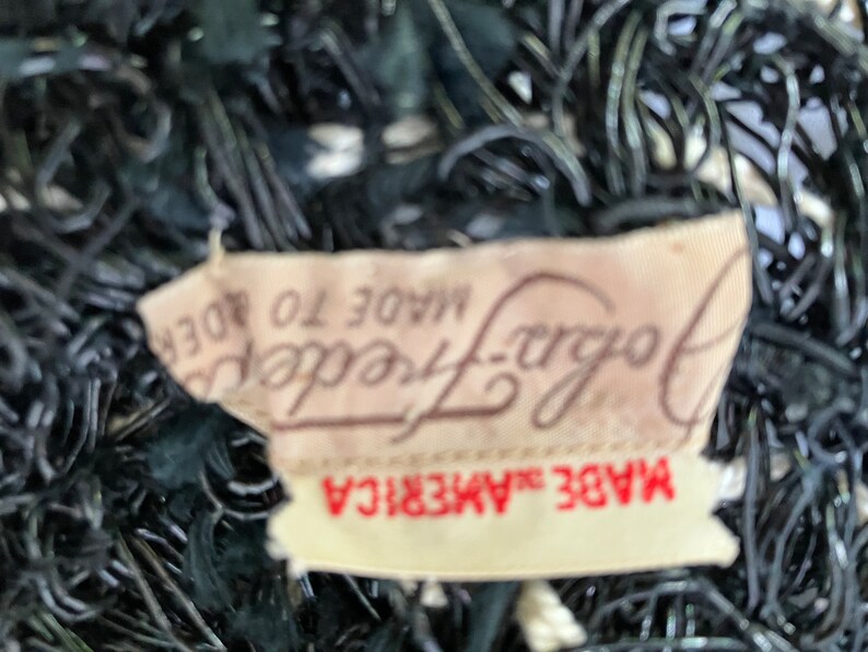 May include: Close-up of a vintage black hat with a woven design. A tag reads "John Frederics Made in America". The hat has a textured, open weave pattern, with a dark color scheme.
