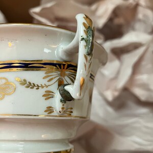May include: A white porcelain teacup with a gold and blue trim. The cup has a handle with a gold and green design.