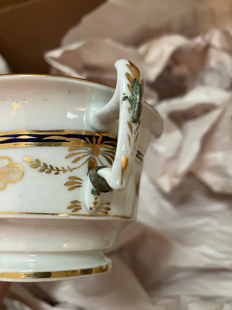 May include: A white porcelain teacup with a gold and blue trim. The cup has a handle with a gold and green design.