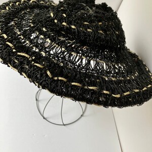 May include: A black and beige woven sun hat with a wide brim. The hat is made of a mesh-like material with a decorative pattern. The hat is displayed on a wire stand against a white background.
