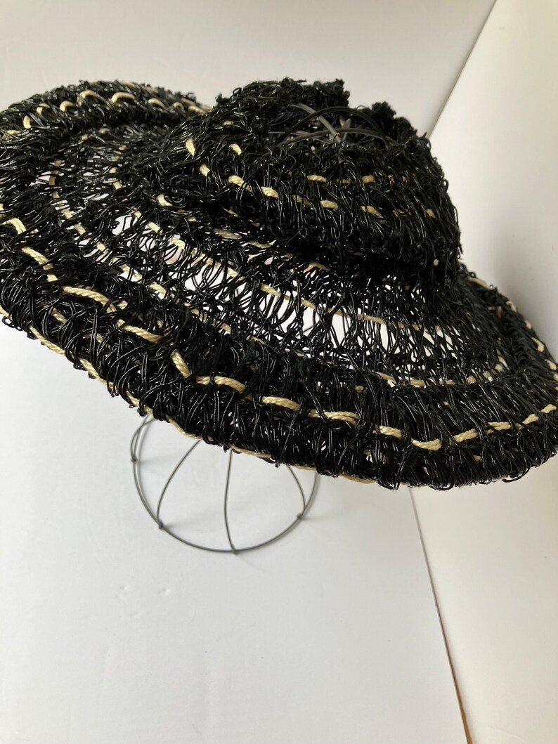 May include: A black and beige woven sun hat with a wide brim. The hat is made of a mesh-like material with a decorative pattern. The hat is displayed on a wire stand against a white background.