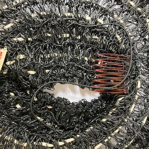 May include: Close-up of a black and white woven hat with a comb and a tag that reads "John Frederics Made in America". The hat has a circular design with a hole in the center.