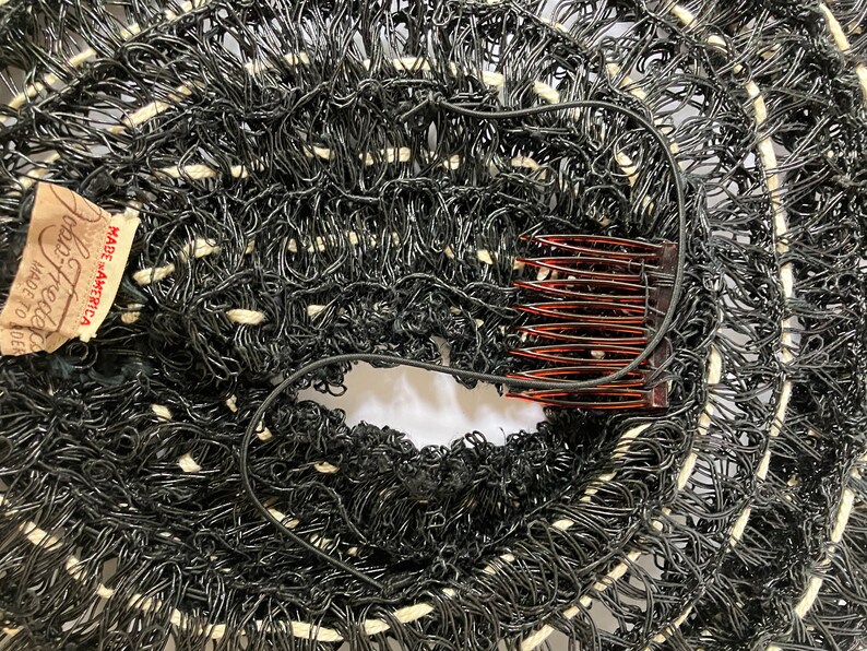 May include: Close-up of a black and white woven hat with a comb and a tag that reads "John Frederics Made in America". The hat has a circular design with a hole in the center.
