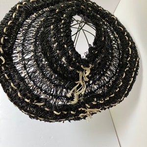 May include: A black and beige woven lampshade with a circular design. The lampshade is made of a dark material with beige accents and a metal frame. The woven pattern creates a textured appearance. The lampshade is designed to diffuse light.