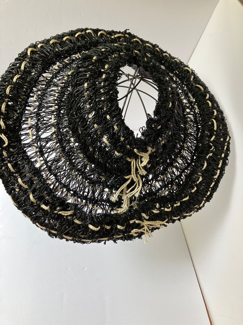 May include: A black and beige woven lampshade with a circular design. The lampshade is made of a dark material with beige accents and a metal frame. The woven pattern creates a textured appearance. The lampshade is designed to diffuse light.