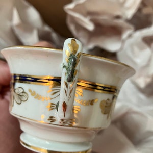 May include: A white porcelain teacup with a gold and blue trim. The cup has a floral design and a gold handle with a green and gold accent.