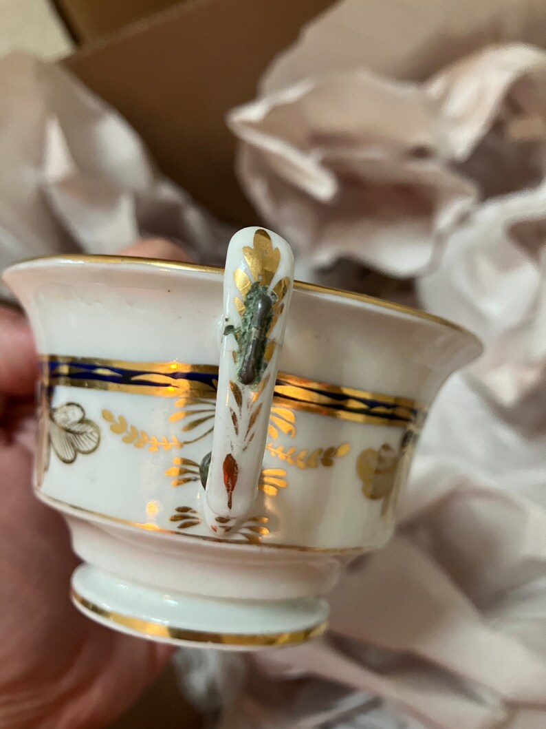 May include: A white porcelain teacup with a gold and blue trim. The cup has a floral design and a gold handle with a green and gold accent.