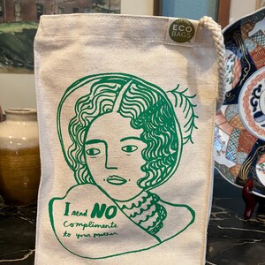 May include: A beige canvas tote bag with a green illustration of a woman's face and the text "I send NO compliments to your mother." The bag has a rope handle and a small "ECO BAGS" tag.