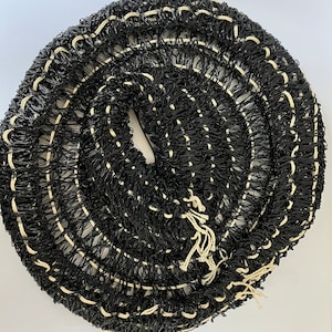 May include: A coiled roll of black netting with white stitching. The netting is tightly woven, creating a textured surface. The roll is centered and takes up most of the frame, with a white background.