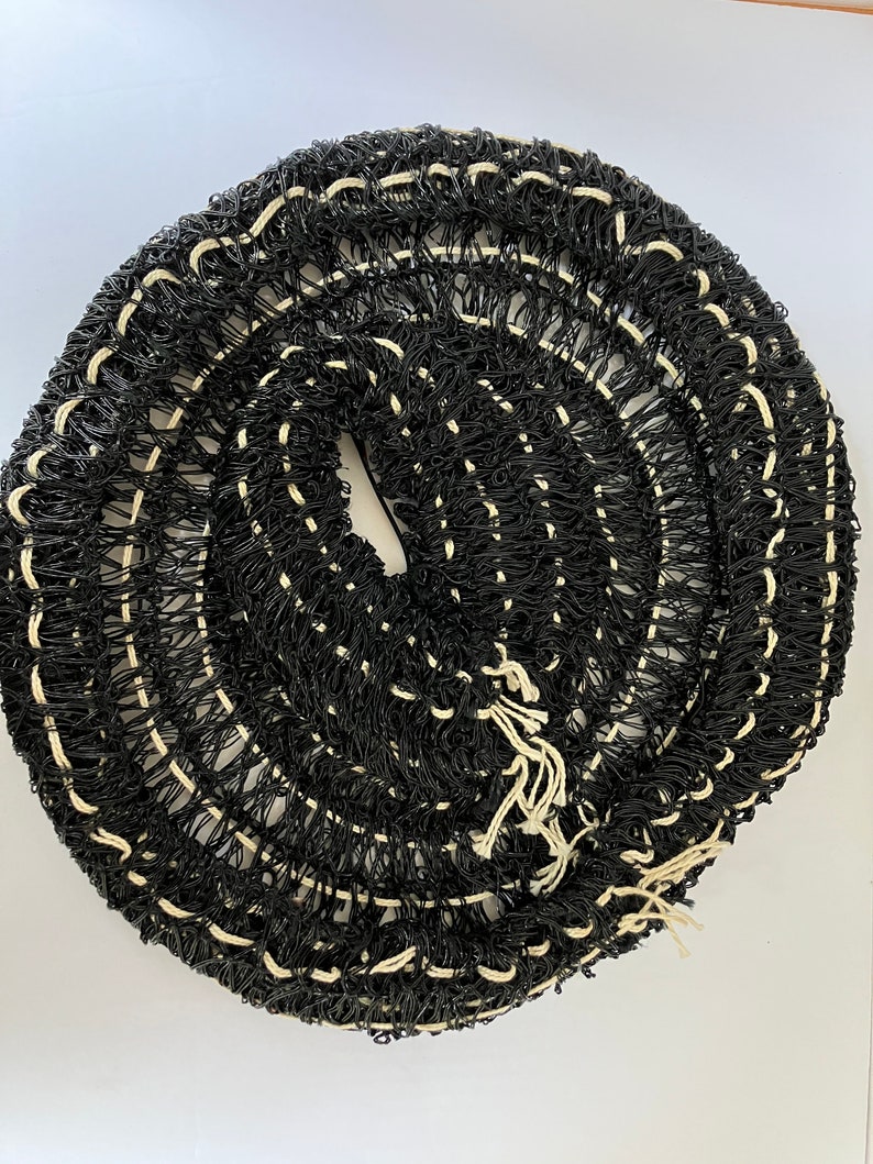 May include: A coiled roll of black netting with white stitching. The netting is tightly woven, creating a textured surface. The roll is centered and takes up most of the frame, with a white background.