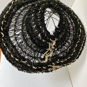 May include: A black and cream-colored woven basket with a circular shape. The basket is made of a dark material with cream-colored accents and a metal frame. The basket has a unique, open weave design.