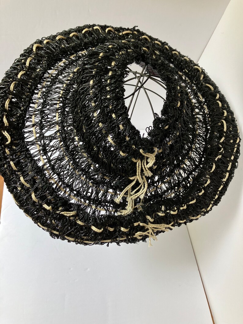 May include: A black and cream-colored woven basket with a circular shape. The basket is made of a dark material with cream-colored accents and a metal frame. The basket has a unique, open weave design.