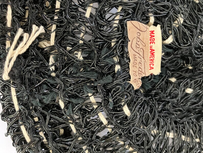 May include: Close-up of a woven textile with a complex pattern of black and off-white threads. The texture is dense and irregular, with a mix of looped and straight strands. A tag reads "John Freeda MADE TO ORDER MADE IN AMERICA."