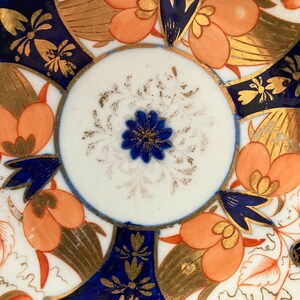 May include: A vintage porcelain saucer with a blue and gold floral pattern. The center of the saucer features a white circle with a blue flower and gold accents.