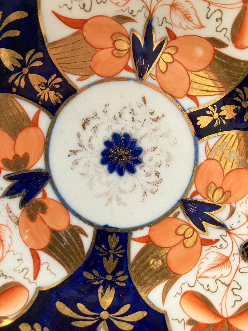 May include: A vintage porcelain saucer with a blue and gold floral pattern. The center of the saucer features a white circle with a blue flower and gold accents.