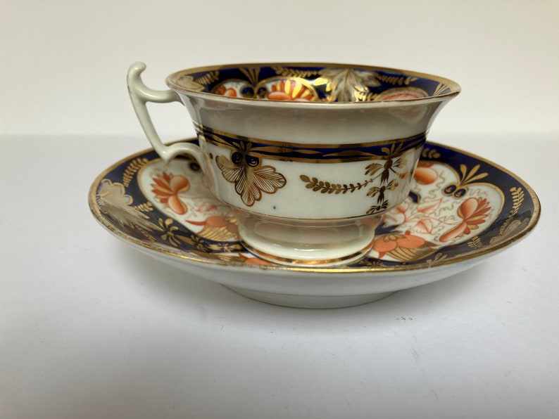 May include: A vintage teacup and saucer set with a blue and gold floral pattern. The cup has a delicate handle and a wide, shallow bowl. The saucer has a matching design and a scalloped edge.