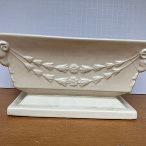 May include: Cream-colored rectangular planter with a pedestal base. The planter features a floral garland design and decorative scrollwork on the ends. A classic piece for displaying plants or flowers.